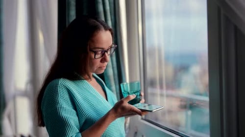 Woman Using Phone and Drinking Water by Window