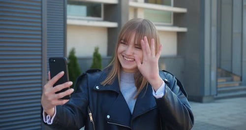 Cheerful Attractive Blonde Woman Talking By Videocall on Smartphone and Waving at Camera in the City