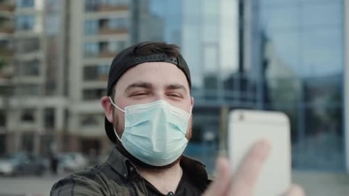 Glücklicher Mann mit medizinischer Maske, der online chattet und der Kamera des Smartphones mit der Hand winkt