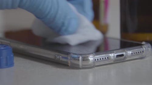 Close-up of Hand in Protective Blue Gloves Rubbing Smartphone Screen with Sanitizer. Unrecognizable