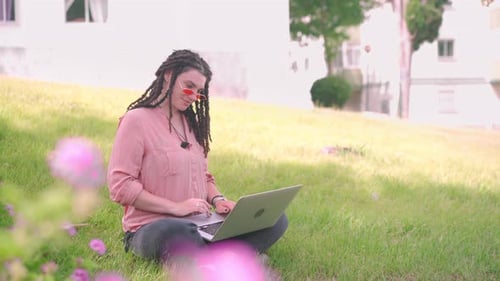 Attractive Woman with Dreadlocks Working at the Laptop Sitting Grass in City Park