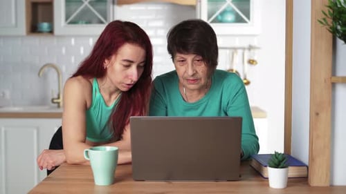 Young Girl Explains to Her Elderly Mother How to Use a Laptop at Home in Kitchen