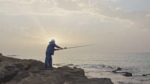 Old fisherman standing on sea side rocks and fishing against the sunset