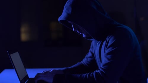 Hooded Figure Typing on Laptop in Dark Room
