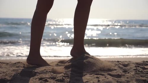 Female Feet Walking at the Sea Beach on a Sunny Day with Waves at Background. Legs of Young Woman