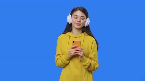 Woman with Headphones Uses Cellphone Against Blue Background