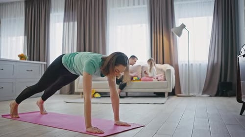 Young Woman Working Out in Living Room Practicing Stretching Exercises While Family Playing Nearby