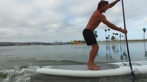 Tracking view of a man paddling his SUP stand-up paddleboard in a lake.