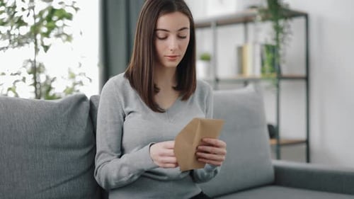 Woman Reads Letter, Smiles on Gray Sofa Indoors