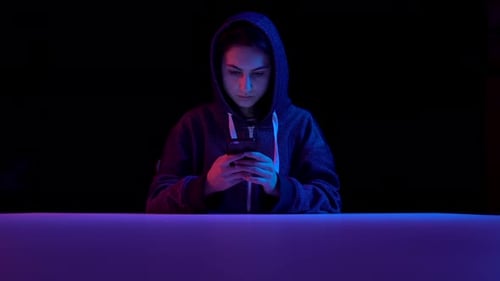 Young Woman in a Hood with a Smartphone. Hacker Makes a Hack Through the Phone. Blue and Red Light