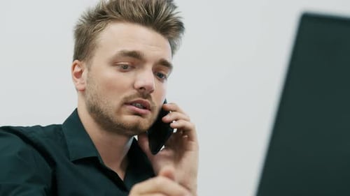 Handsome Young Bearded Man Talking on Mobile Phone and Using Laptop at Office