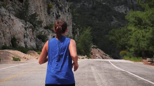 Back follow shot of young woman running on road surrounded by green mountains.