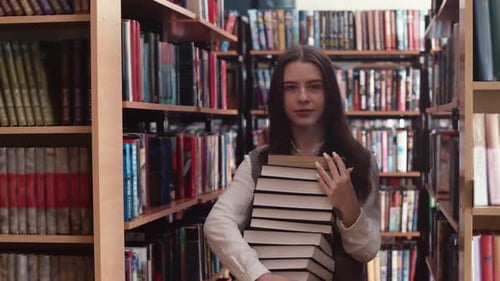 Young Caucasian Female Student Holds Stack of Books in Library Among Shelves with Books Front View