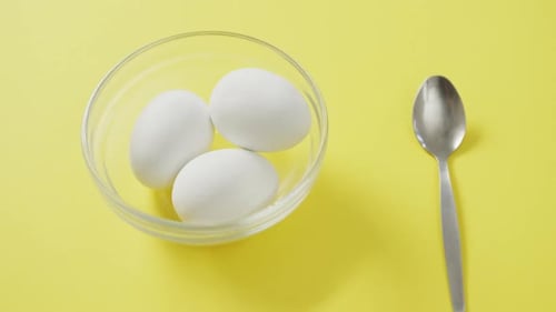 Video of overhead view of glass bowl with eggs and silver spoon on yellow background