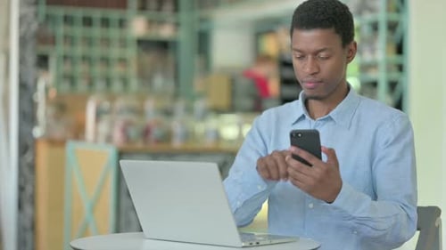 Young African Man Using Smartphone and Laptop in Cafe