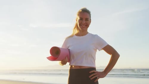 Portrait of caucasian woman holding yoga mat at the beach smiling