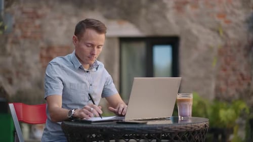 A Man Sitting in a Street Cafe Works Remotely Writing a Pen in a Notebook