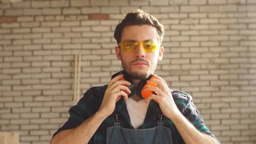 Portrait of a Young Man in a Carpenter's Workshop in Protective Glasses Looking at Camera. Concept