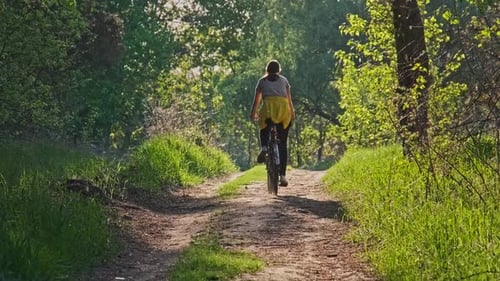 Young Woman on a Bicycle Rides Along Green Forest Path in Sunny Summer Day