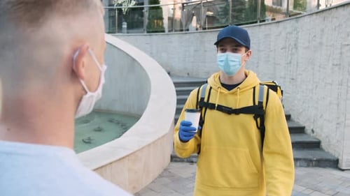 Delivery Man Wearing Thermal Backpack and Medical Mask Delivers a Coffee to a Young Male Customer
