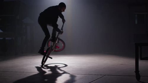 BMX rider doing tricks in dark warehouse.
