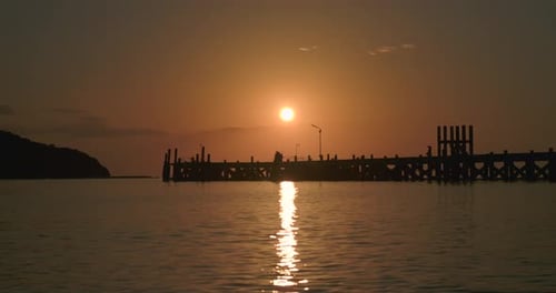 Warm orange sun above sea with reflection on water surface and pier silhouette during sunset
