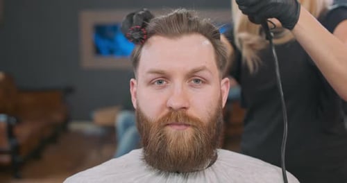 Close Up Head of a Young Bearded Man and Hands of Young Female Barber Who Dries His Hair with a