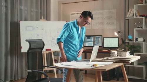 Asian Man Engineer Walk Into The Office Before Looking At Blueprint And Typing On A Laptop