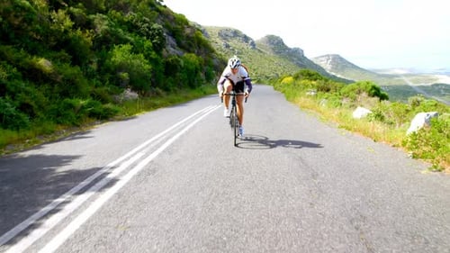 Female cyclist cycling on a countryside road beside a lush mountain