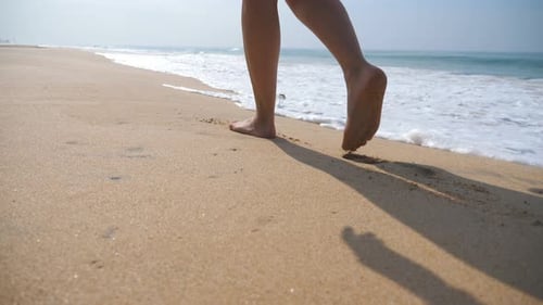 Female Feet Walking on Golden Sand at the Beach with Ocean Waves at Background. Legs of Young Woman