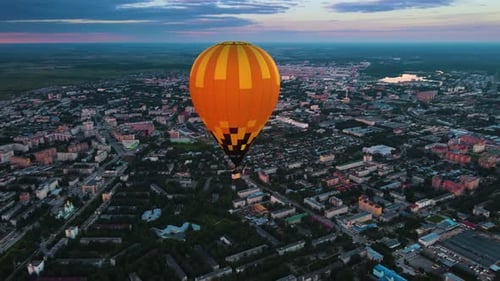 Hot air balloon flies over evening landscape. Bright envelope glows against city