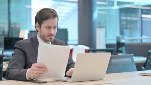 Businessman Reviews Documents at Desk with Laptop