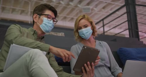Asian man and caucasian woman wearing face masks discussing over a digital tablet at office