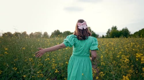 Back View of Rural Woman in Turquose Dress Walks Among Blooming Canola Yellow Flowers in Field