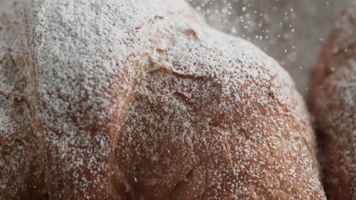 Camera follows sprinkling powered sugar over croissant. Slow Motion.