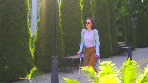 Positive Beautiful Stylish Young Woman with an Injury Walking on Crutches Through the Park in the