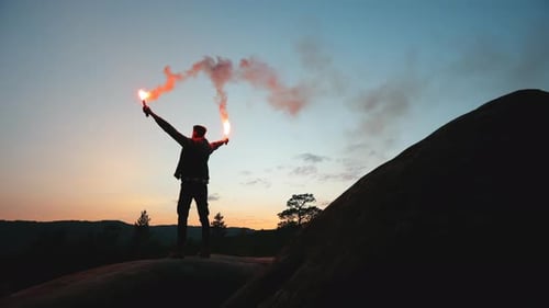 A Man is Standing on the Edge of a Cliff and Waving Two Flares in the Night