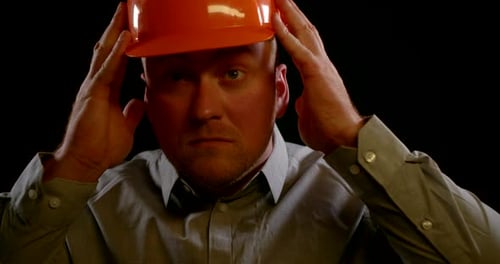 Adult Bald Man Is Putting Off Orange Helmet From His Head, Closeup Portrait at Black Background