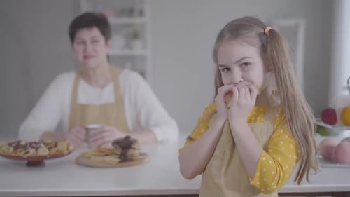 Portrait of Cute Little Girl Eating Pancake with Blurred Caucasian Grandmother Smiling at the