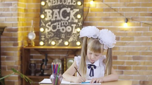 Cute Little Girl Sits at Table and Draws with Colored Pencils on the Background of a School Class