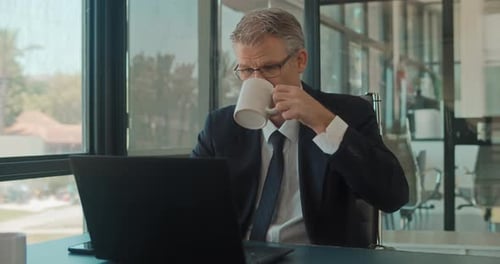 A businessman in formal wear and glasses having a drink while working on a laptop at the office