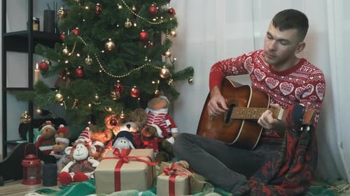 Handsome man in red winter sweater is sitting on plaid near Christmas tree and playing guitar