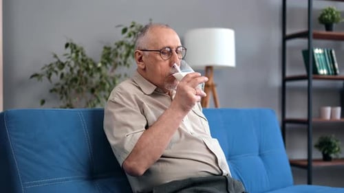 Pensive Elderly Man Drinking Fresh Milk Glass Smiling Relaxing on Comfortable Blue Couch at Home