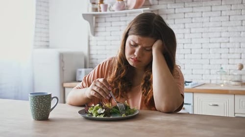 Frau in Übergröße sitzt am Tisch und fühlt sich unzufrieden mit der Ernährung und möchte keinen Salat essen