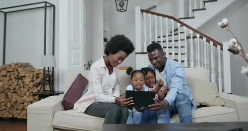 Padres afroamericanos con hijas pequeñas sentados juntos en el sofá y usando una tableta