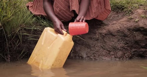 Hands collect water in plastic container from a dirty source in Uganda, Africa.