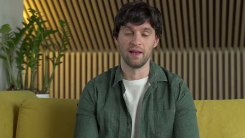 A Happy Man in a Green Shirt Looks at the Camera Talks Via Video Link with Friends Sits at Home on a