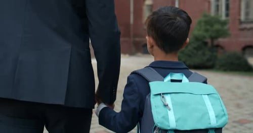 Vista de cerca de un niño con una bolsa tomando la mano de su padre y caminando hacia la escuela. Persona masculina en