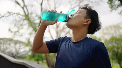4K Asian man drinking water from a bottle while jogging at public park in the morning.