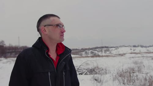 An Adult Man with Glasses Looks Thoughtfully Into the Distance Standing on the Street in Winter in